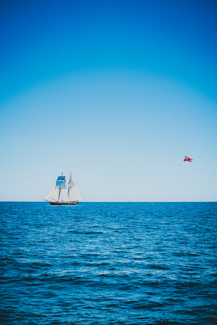 A tall ship on open blue water with a small red plane overhead